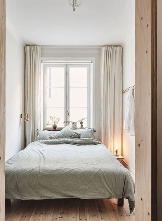 a neutral narrow bedroom with a bed with olive bedding, a rack and a nightstand plus a wall lamp