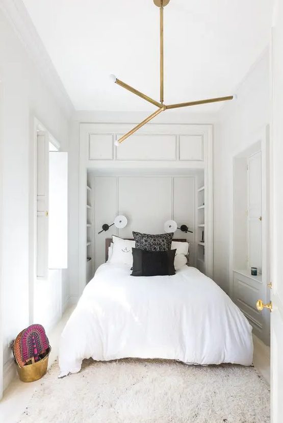 a neutral narrow bedroom with built-in shelves at the headboard, a bed with black and white bedding, a chandelier and some bright decor