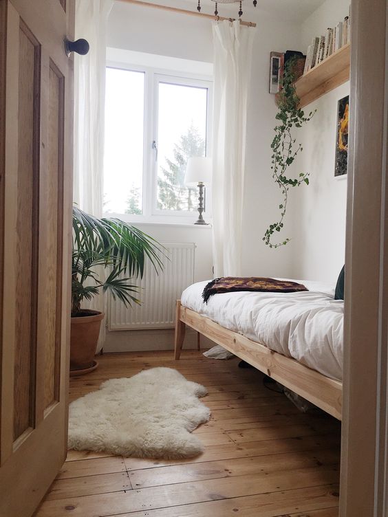a small and narrow bedroom with stained furniture, neutral textiles, potted plants and some decor on the wall