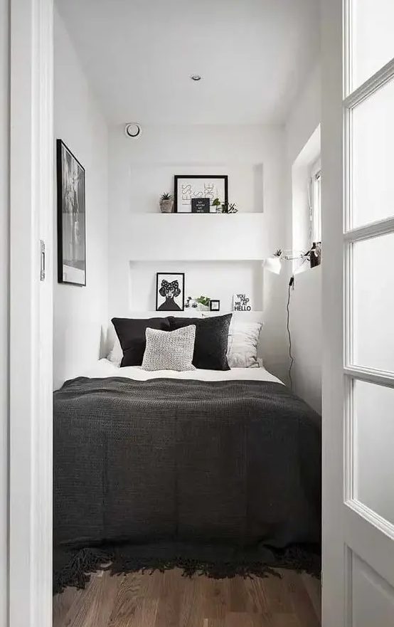 a tiny bedroom in white, with only a bed squuzed in, black and white bedding, niche shelves with decor, an artwork and a sconce
