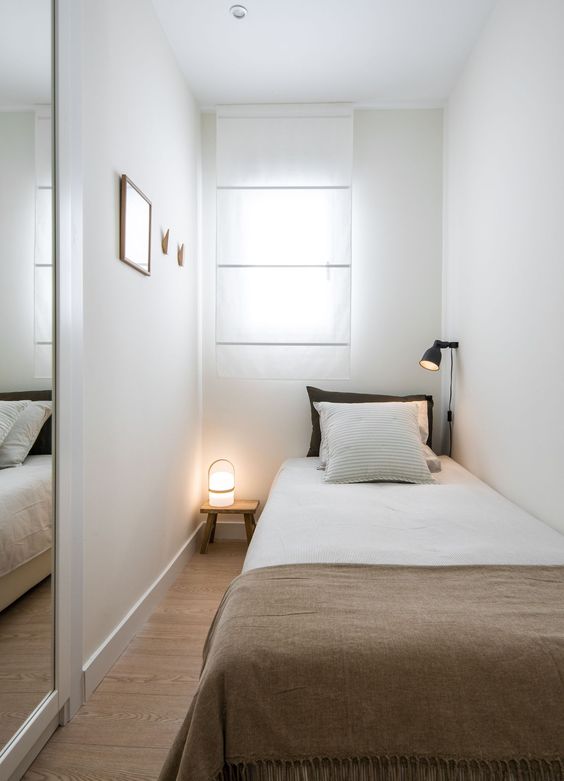 a tiny narrow bedroom with a window, a single bed, neutral bedding, a wall lamp and a small nightstand