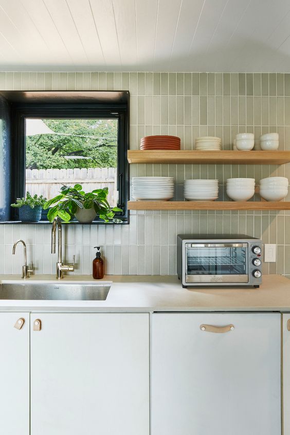 a white kitchen with lower cabinets and shelves and a light green stacked tile backsplash is a cute and welcoming space