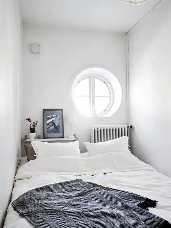 a white narrow bedroom with a porthole window, a bed with neutral bedding, a narrow nightstand at the headboard and a radiator