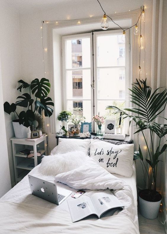 a white narrow bedroom with a small bed, a nightstand with storage, potted plants, decor and lights over the bed