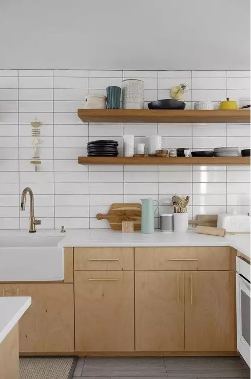 an airy Scandinavian kitchen with light colored wooden cabinets and shelves, a white stacked tile wall and brass fixtures