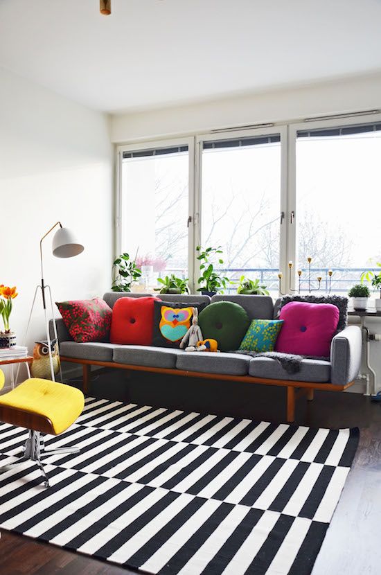 a-pretty-modern-living-room-with-a-glazed-wall-a-grey-sofa-with-colorful-pillows-a-striped-rug-a-yellow-chair-a-floor-lamp-and-some-greenery.jpg