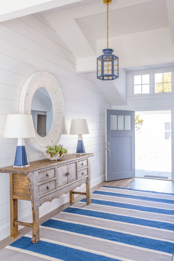 an-amazing-coastal-entryway-with-white-planked-walls-a-blue-and-grey-striped-rug-a-wooden-console-table-and-blue-touches.png