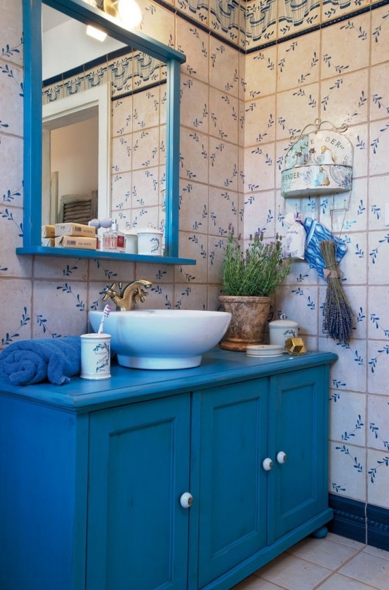 a bright Provencal bathroom with floral wallpaper, a bold blue vanity and a mirror in a blue frame, a bowl sink and a vintage faucet, lavender and blooms