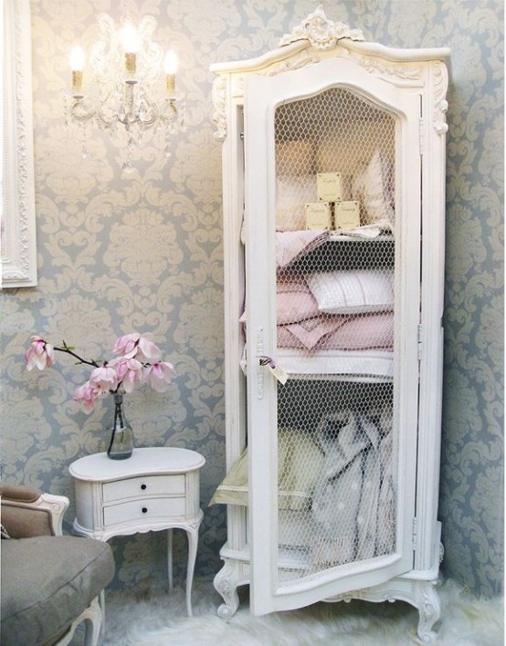 a refined Provence bathroom with printed wallpaper, a refined and chic storage unit with a chekcen wire door and towels and other stuff inside