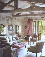 a rustic Provence living room with stone walls, wooden beams, refined vintage furniture, pink curtains and a pink ottoman as a table