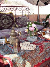 a colorful Moroccan terrace with a metal bench with purple upholstery and pillows, a table covered with a red printed blanket, Moroccan teaware and blooms