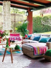 a colorful boho terrace with wicker furniture, bold blankets and pillows, a rattan screen, potted greenery and blooms
