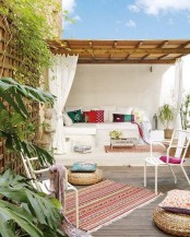 a contrasting and eye-catchy boho terrace with a built-in sofa, colorful pillows, blankets and a rugs, white chairs, potted greenery and blooms