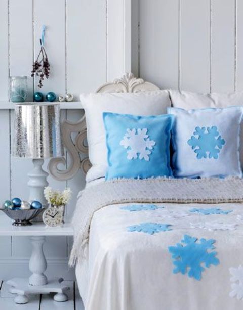 a blue and white Christmas bedroom with snowflake pillows, blue and white ornaments, a silver lamp and a nightstand