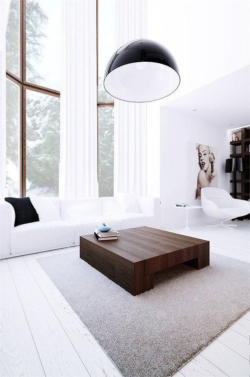 a white minimalist living room with double height windows, a white sofa, a dark stained coffee table and a pendant lamp
