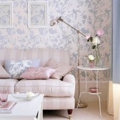 a delicate pastel living room with printed wallpaper, a striped sofa, pastel pillows, a side table with a lamp and white and pink blooms and a low coffee table