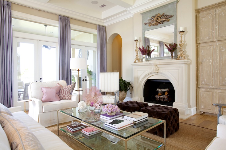 a vintage inspired feminine living room with a large fireplace, white seating furniture, lilac curtains, a glass coffee table and pink pillows