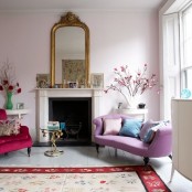 a lovely girlish living room with vintage touches, with light pink walls, a fireplace, a lilac loveseat and a hot red chair, touches of gold and some blooms