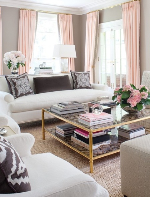 a chic feminine living room with grey walls, grey and neutral seating furniture, pink curtains and a rug, printed pillows and blooms
