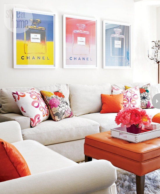 a bright feminine living room with neutral walls and a sectional sofa, colorful pillows, an orange leather ottoman and a glam print gallery wall