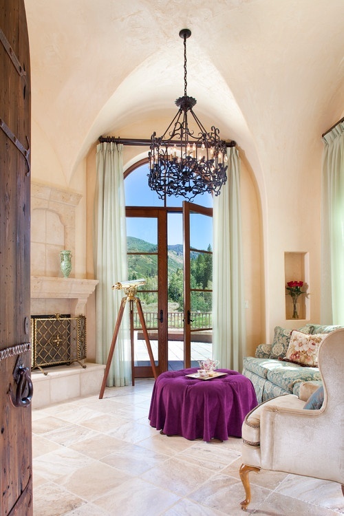 a gorgeous living room with arched windows, a fireplace, a vintage chandelier, a green sofa and a neutral chair, a fuchsia pouf is wow