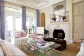 a glam vintage-inspired neutral living room with a fireplace, white seating furniture, lilac curtains, touches of pink and a glass coffee table