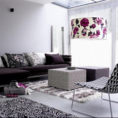 a white living room with a deep purple sofa, a purple and a printed pouf, a printed rug and a faux fur one, a geo printed chair and a chic floral lampshade