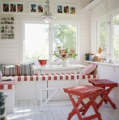 a white Scandinavian sunroom with a built-in bench with striped upholstery, red stools and a dining table and a gallery wall