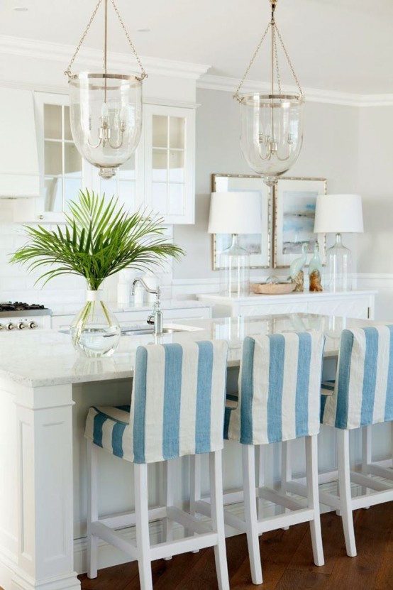 a beautiful tropical white kitchen with shaker style cabinets, white stone countertops, cool striped stools and greenery and elegant glass pendant lamps