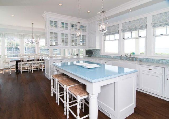 a coastal kitchen with a blue tile backsplash, white shaker style cabinets, a blue countertop, woven stools and glass pendant lamps