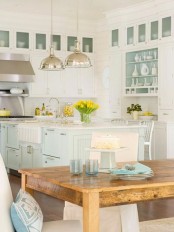 a pretty and delicate beach kitchen with white cabinets, a very light aqua kitchen island, shiny metal pendant lamps and a wooden table with aqua accessories