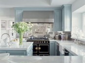 an elegant light blue kitchen with shaker cabinets, white stone coutnertops, a black and gold cooker and a shiny silver tile backsplash