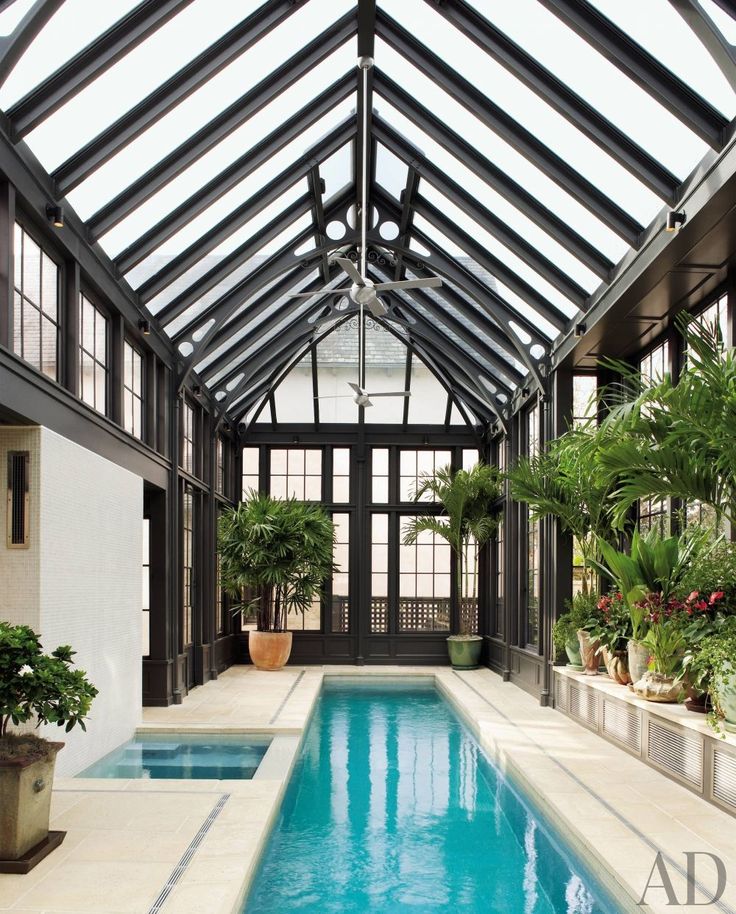 a large glazed pavilion with black framing, a long and narrow pool, a stone deck, a hot tub and potted plants and blooms by its side