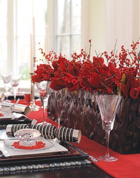 a bright Christmas tablescape with a red table runner, a black planter with red berries and poinsettias, elegant black square plates and chic glasses