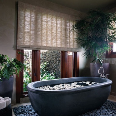 a tropical bathroom with a stone tub placed into pebbles, potted greenery and a large window that goes to a private garden