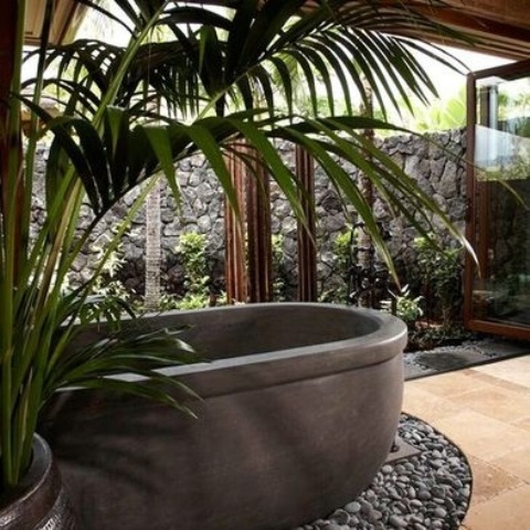 a stylish indoor outdoor tropical bathroom with a large folding door to open it to the garden and a stone tub and potted plants