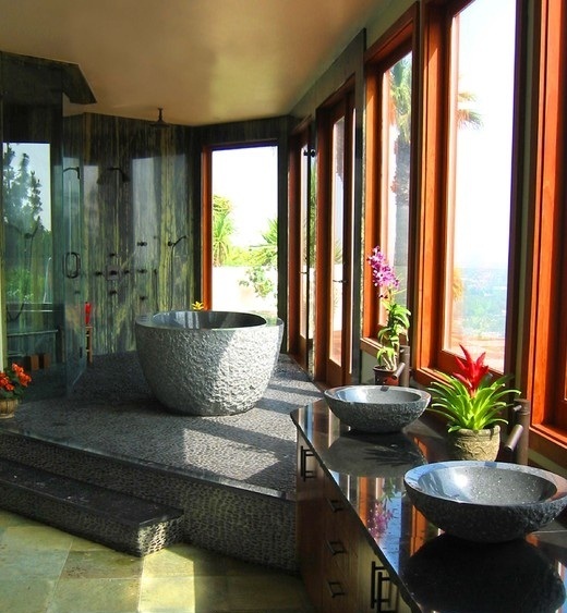 a modern tropical bathroom with all glazed walls, a stone platform, a stone tub and a shower, stone sinks and potted plants