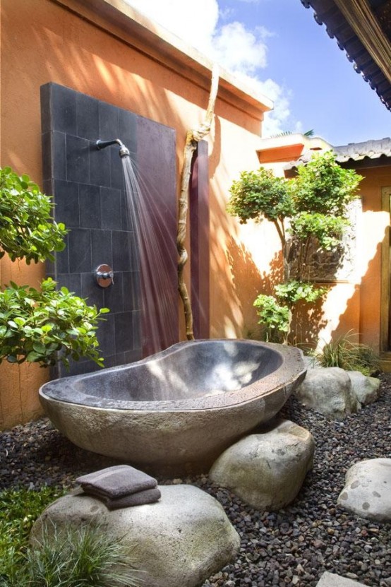 an outdoor tropical bathroom with a tile wall, a stone tub, rocks, pebbles and potted greenery and trees