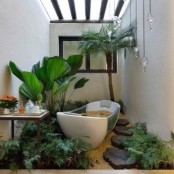 a tropicla bathroom in neutrals, with wooden beams, a tub placed in pebbles, statement plants growing around and pendant lamps