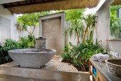 an outdoor-indoor tropical bathroom clad with neutral stone, with a stone tub and tropical plants growing around plus stone sinks