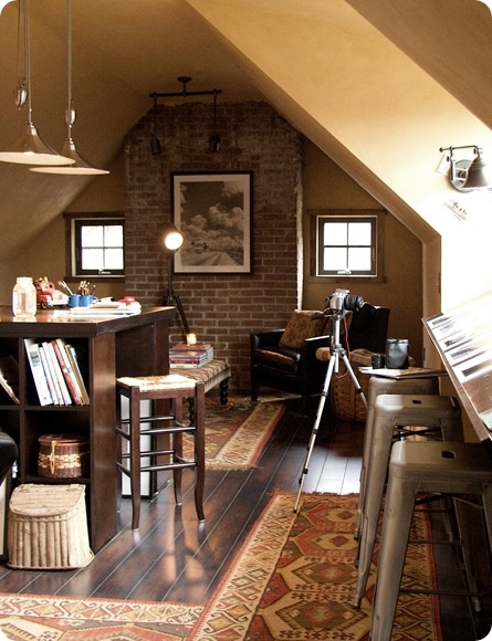 an industrial home office with small windows and a skylight, a dark stained desk, metal and wooden stools, a black leather chair, pendant lamps and printed rugs plus wall sconces