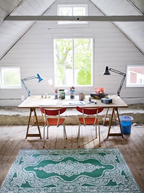 an attic home office with planked walls and a ceiling, a wooden beam and lots of windows, a trestle desk, orange chairs, a printed rug and table lamps