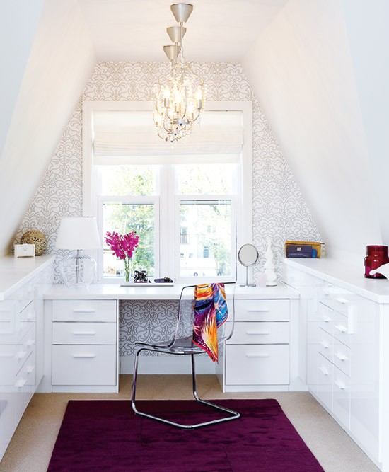 a glam attic home office with two storage units and a built-in desk, an accent wall done with printed wallpaper, chic glam chandeliers, an accent purple rug and a sheer chair