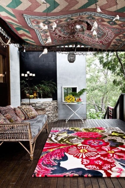 a tropical boho porch with a rattan daybed and a table, super colorful textiles and a blanket over the space plus greenery