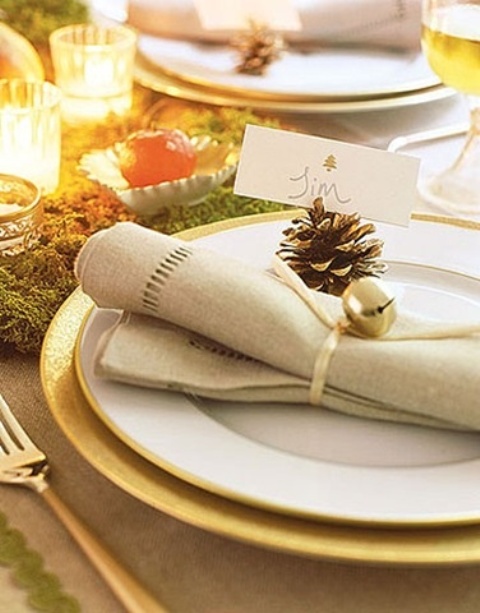 a place setting with a gold charger, a white plate, a neutral napkin with a pinecone and a bell is very chic and Christmassy
