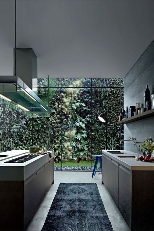 a beautiful minimalist kitchen with sleek dark cabinets, white stone countertops, an open shelf, a kitchen island and a glass wall that shows off greenery in a private garden