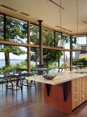 a beautiful modern kitchen and dining space in one, with glazed walls, a hearth, a light-stained kitchen island, a glass table and woven chairs is chic