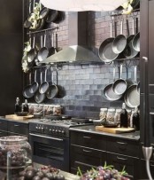 an elegant dark kitchen with tiles, a metal hood, dark cabinets and surfaces, lots of pots hanging on hangers