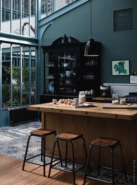 a moody kitchen with dark green walls, refined black furniture, light colored wooden furniture and black pendant lamps
