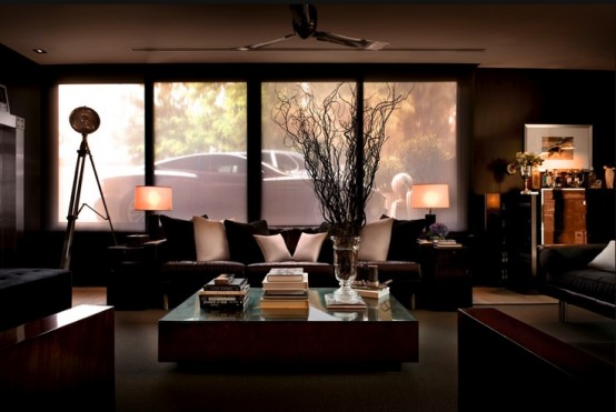 a dark masculine living room with elegant upholstered furniture, a glass coffee table, lamps and a large window